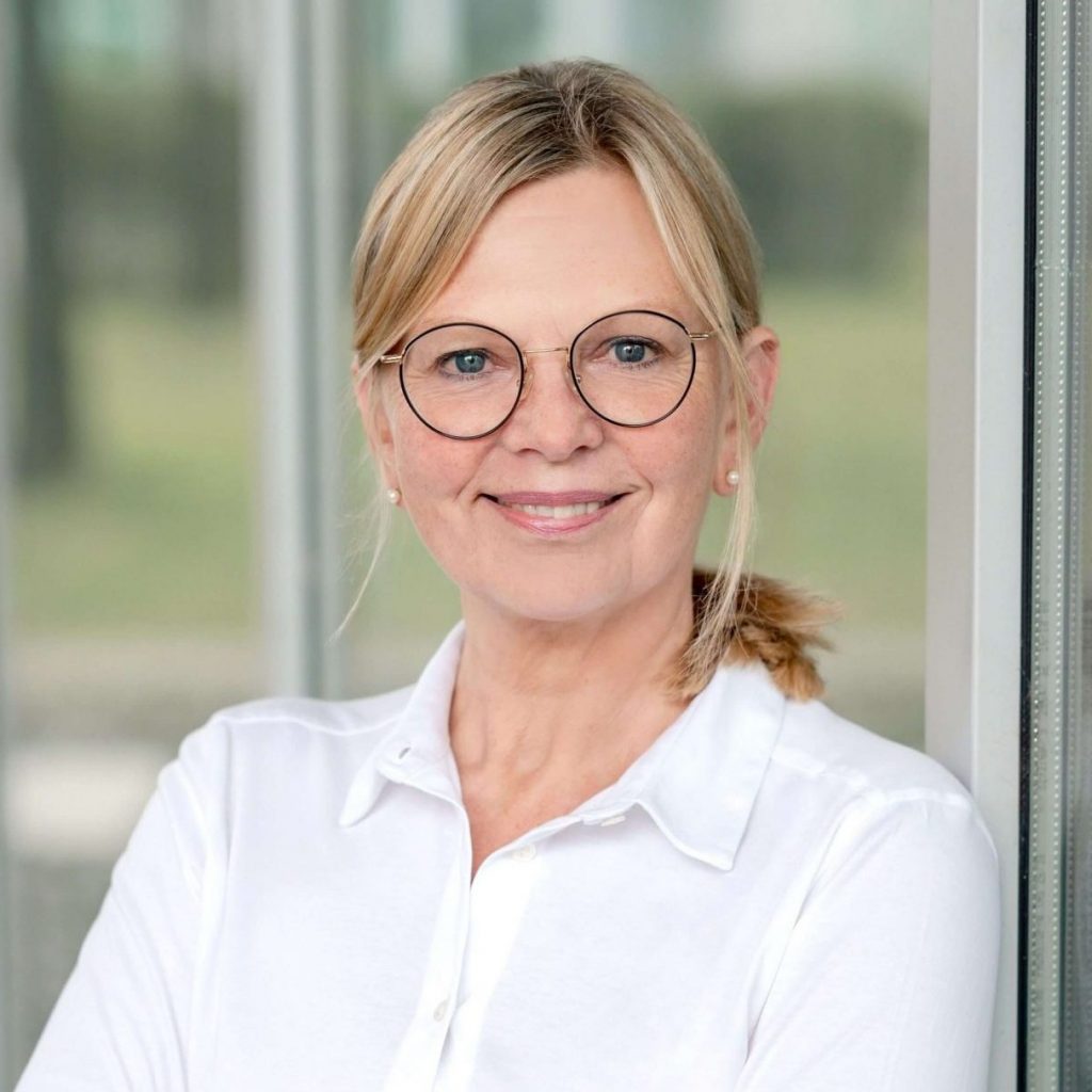 Sie sehen das Portraitfoto einer lächelnden Frau mittleren Alters mit blonden, zum Pferdeschwanz zusammengebundenen Haaren. Sie trägt eine große ovale Brille mit dünnem dunklen Rahmen und ein weißes Poloshirt.