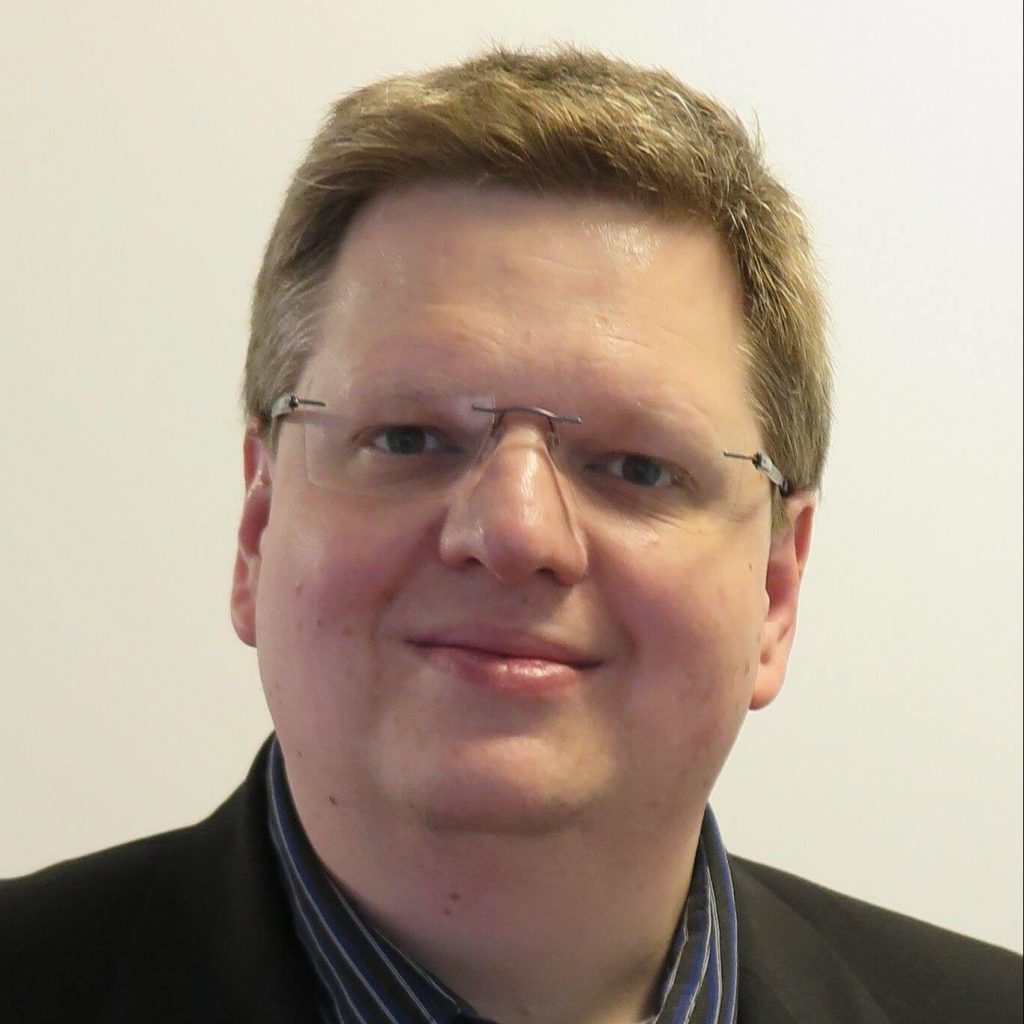 Sie sehen das Gesicht eines Mannes mittleren Alters. Die Haare sind blond und kurz, er trägt eine Rahmenlose eckige Brille mit silbernen Bügeln.