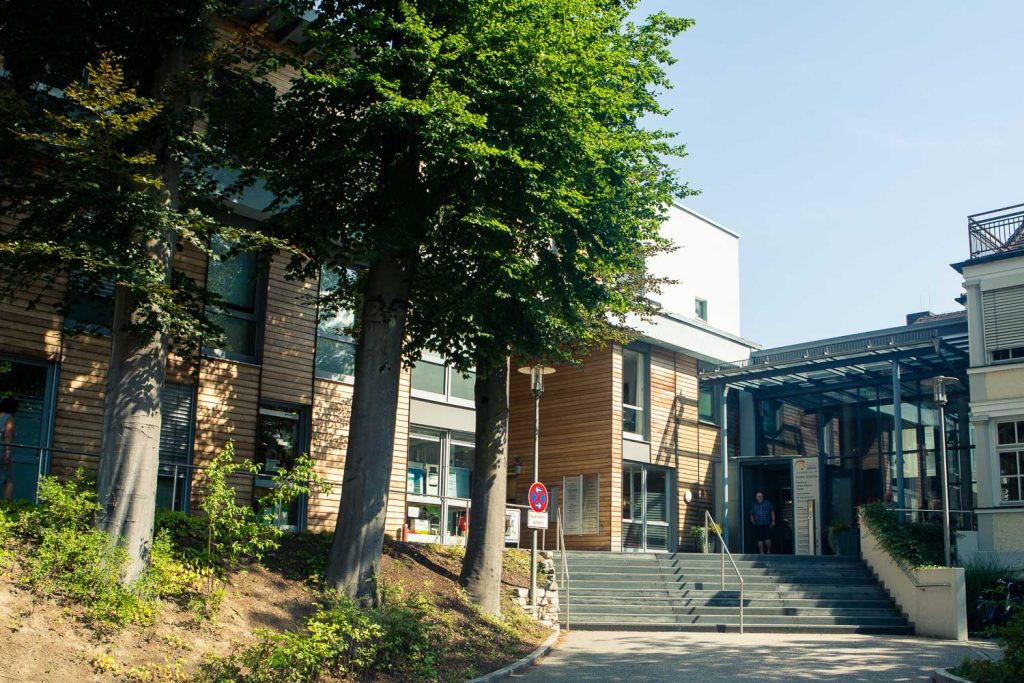 Sie sehen die Außenasicht eines Gebäudes, der Großteil der Fassade ist aus hellem Holz und Glaselementen. Auf der linken Seite sind Bäume davor, rechts führt eine Breite Treppe zum Gebäude hinauf.