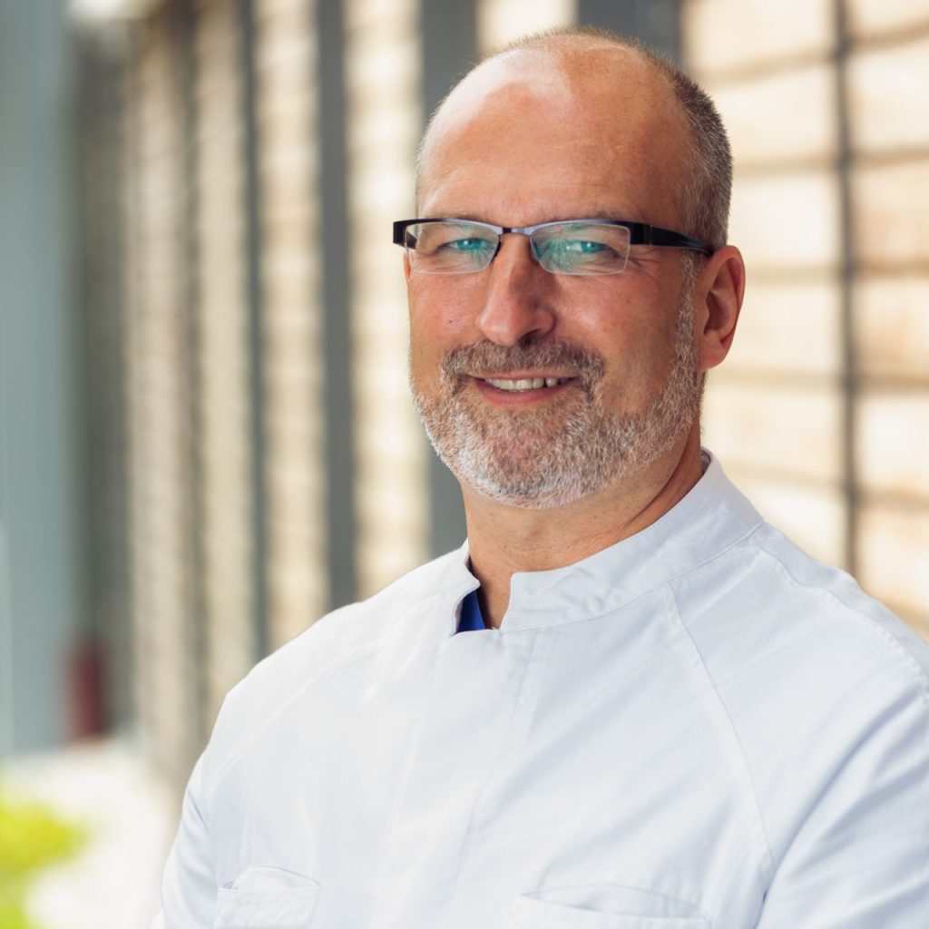 Sie sehen ein Portraitfoto eines lächelnden Mannes mittleren Alters in weißem Arztkittel. Die Haare sind kurz, oben deutlich rückläufig und ebenso weiß-grau wie sein Bart. Er trägt eine eckige schmale Brille mit dicken schwarzen Bügeln.