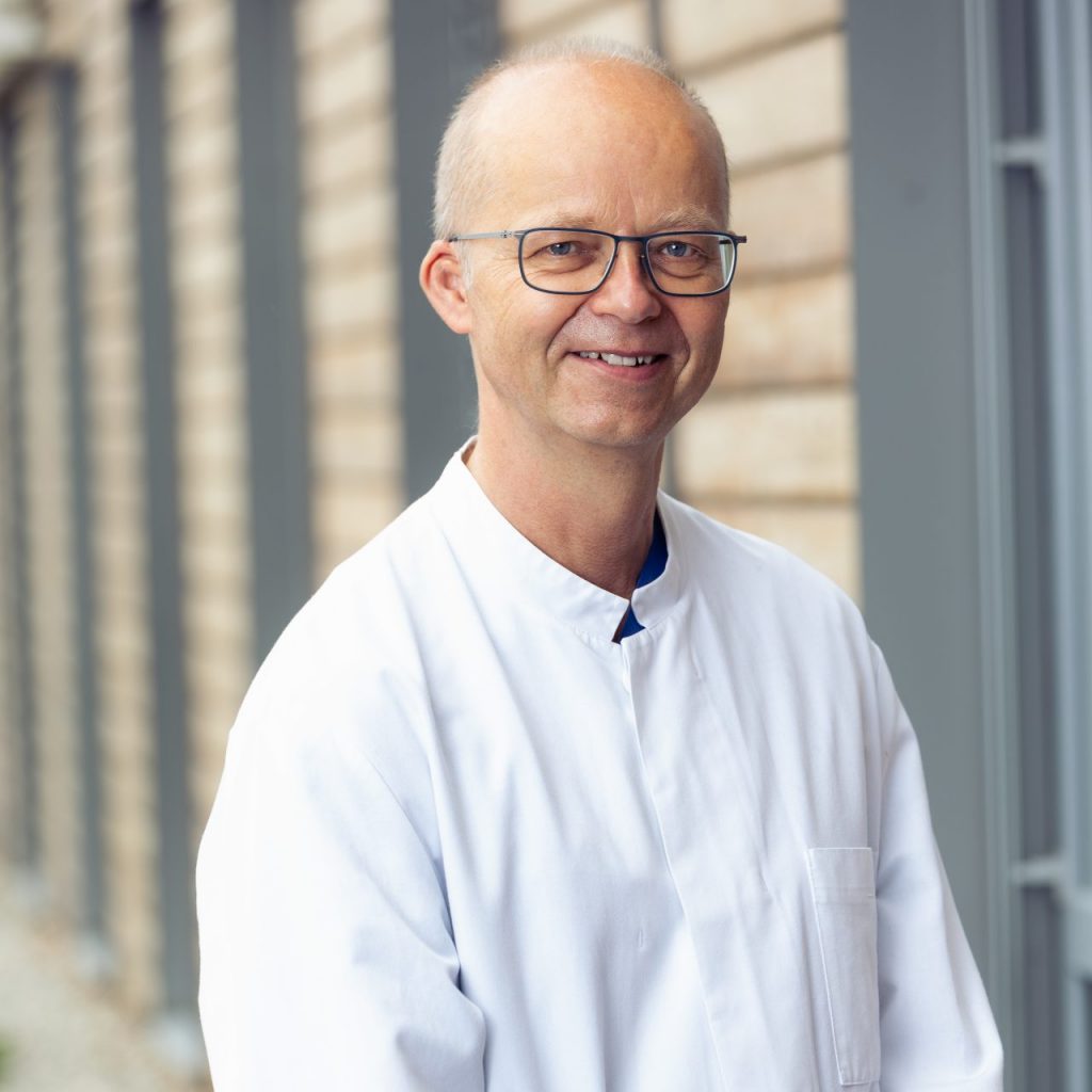 Sie sehen ein Portraitfoto eines Mannes mittleren Alters. Er trägt einen weißen Arztkittel und lächelt freundlichen in die Kamera. Seine Haare sind weiß kurz und dünn. Er trägt eine eckige Brille mit dünnem schwarzem Gestell.