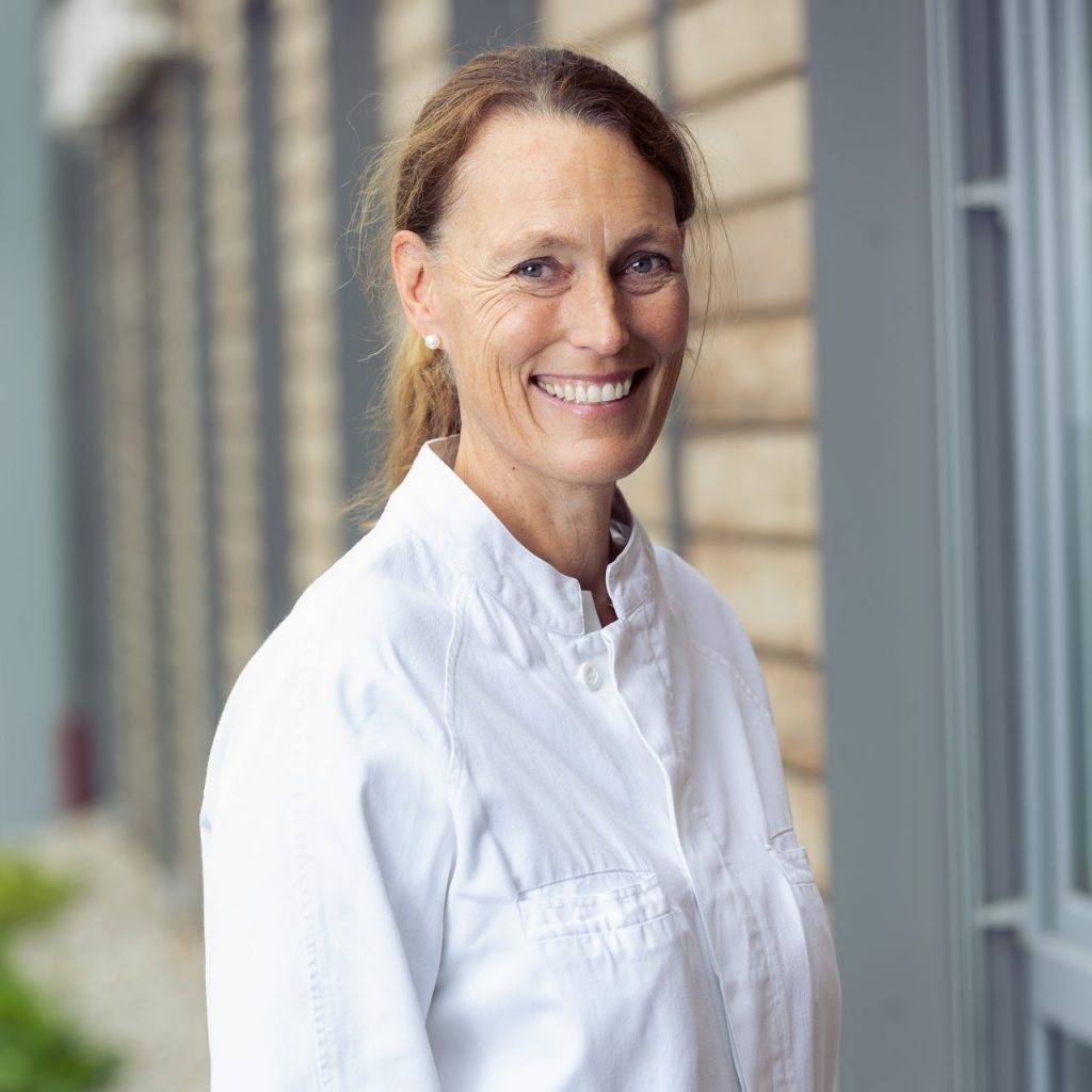 Sie sehen eine freundlich lachende Frau mittleren Alters in weißem Arztkittel. Sie trägt ihre rot-braunen Haare zu einem Pferdeschwanz gebunden.