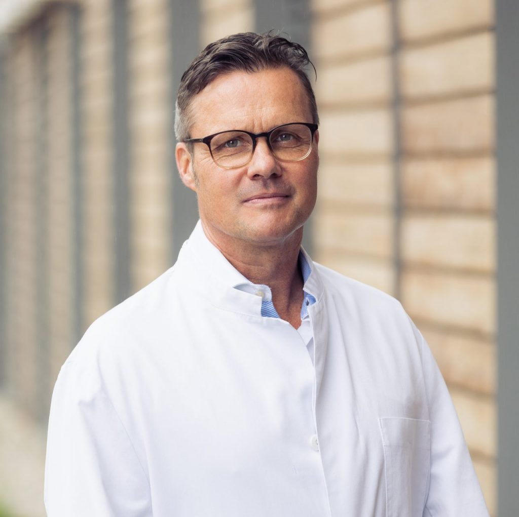 Sie sehen ein Portraitfoto eines Mannes mittleren Alters in weißem Arztkittel. Die lockigen braunen, an den Schläfen ergrauenden Haare sind oben zurückgegelt. Er trägt eine rundliche Brille mit dunkler Fassung.