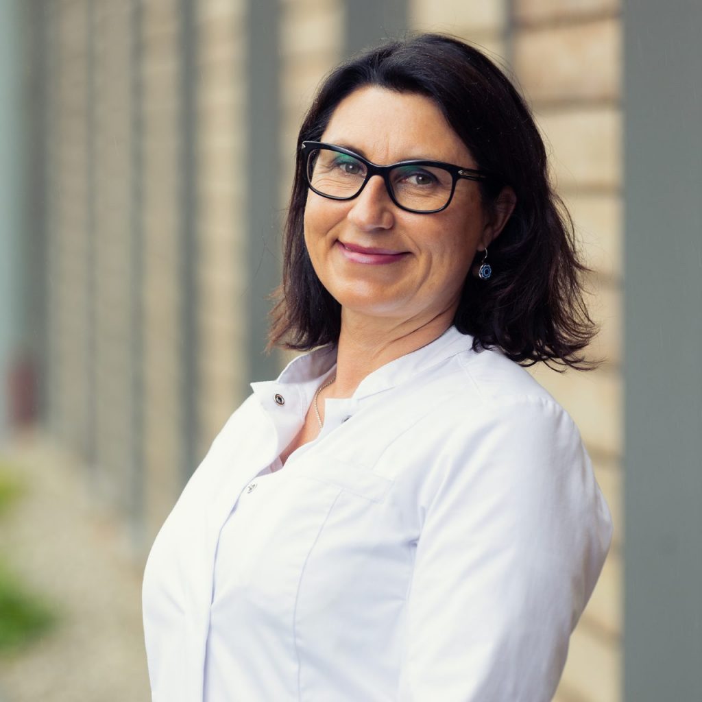 Sie sehen das Portraitfoto eines lächelnden Frau mittleren Alters in einem weißen Arztkittel. Das dunkle Haar ist voll und schulterlang. Sie trägt eine große eckige Brille mit schwarzem Gestell.
