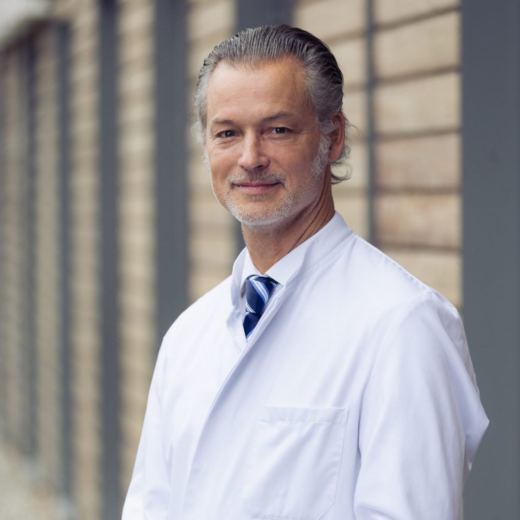 SIe sehen das Portraitfoto eine Mannes mittleren Alters in weißem Arztkittel mit Hemd und blauer Krawatte darunter. Das Haar ist grau weiß und zurückgelegt und gekämmt, der Bart weiß. Er