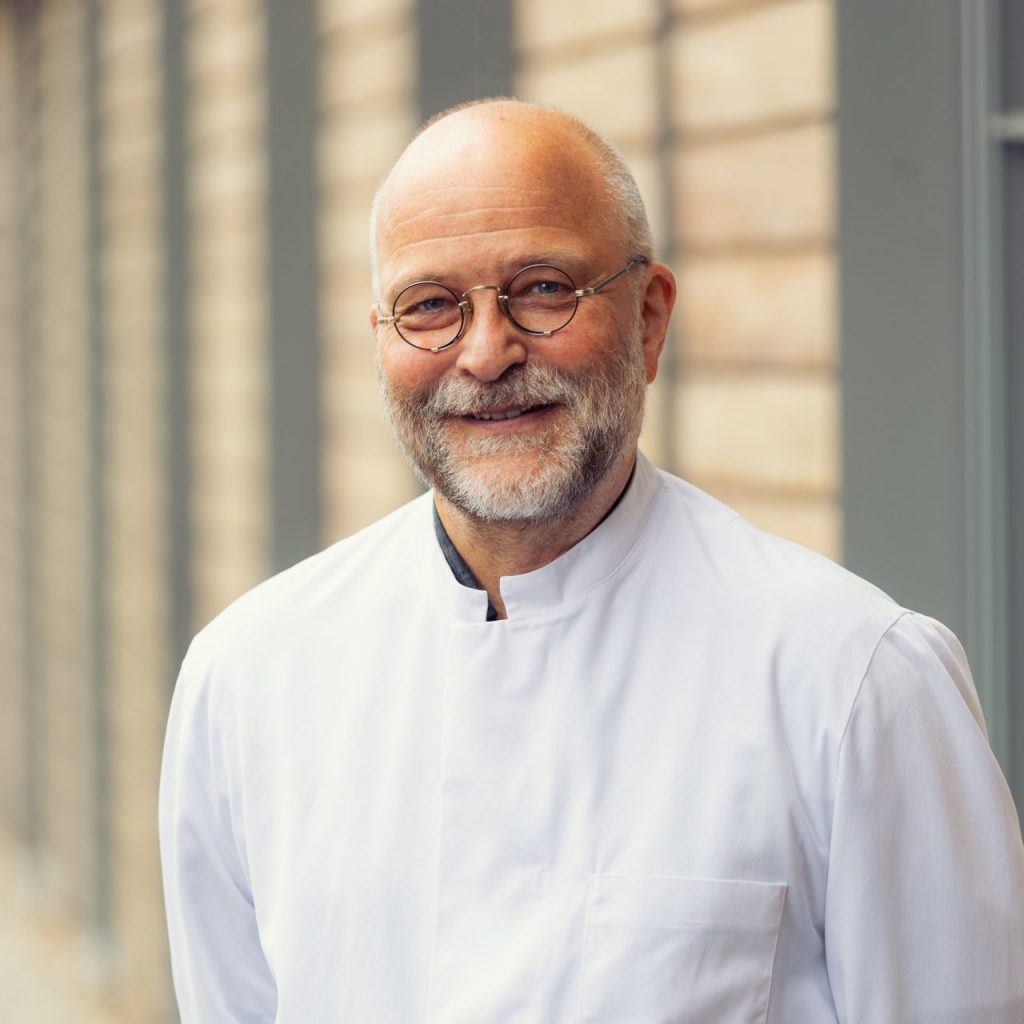 Sie sehen ein Portraitfoto eines lächelnden Mannes mittleren Alters in weißem Arztkittel. Die Haare sind kruz weiß und oben deutlich zurückgegangen, der volle Bart ist ebenfalls weiß mit grauen Stellen. Er trägt eine runde Brille mit silbernem Gestell.