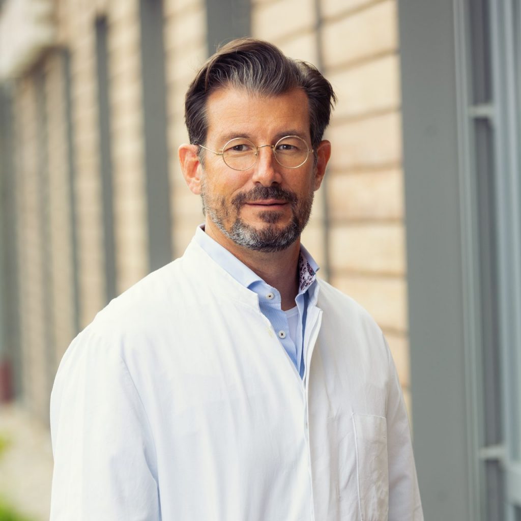 Sie sehen ein Portraitfoto eines Mannes mittleren Alters. Er hat dunkles zur linken Seite gewelltes Haar und Bart. Er trägt eine runde, fein silbern gerahmte Brille und ein hellblaues Hemd unter einem weißen Arztkittel.