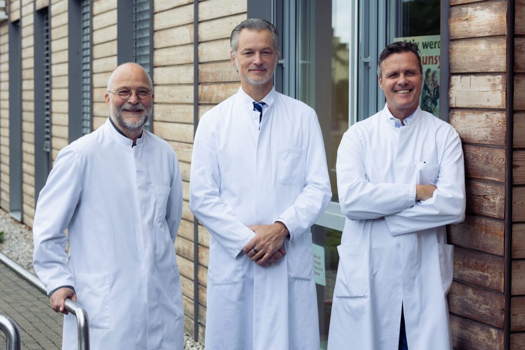 Sie sehen ein Gruppenfoto dreier Männer mittleren Alters in weißen Arztkitteln, die freundlich in die Kamera blicken.