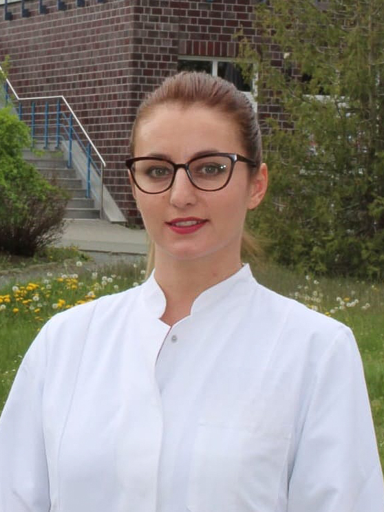 Sie sehen eine lächelnde junge Frau in weißem Arztkittel. Das braune Haar ist zu einem Pferdeschwanz gebunden und sie trägt eine geschwungene Brille mit dunklem Gestell.