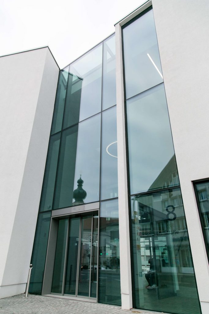 Sie sehen die Außenansicht eines dreistöckigen Gebäudes. Die Wände sind in hellem grau, die Mitte ist mit deckenhohen Fenstern aus grünlichem Glas gestaltet und zentral in diesem Beriech sehen Sie im Erdgeschoss eine Schiebetür.