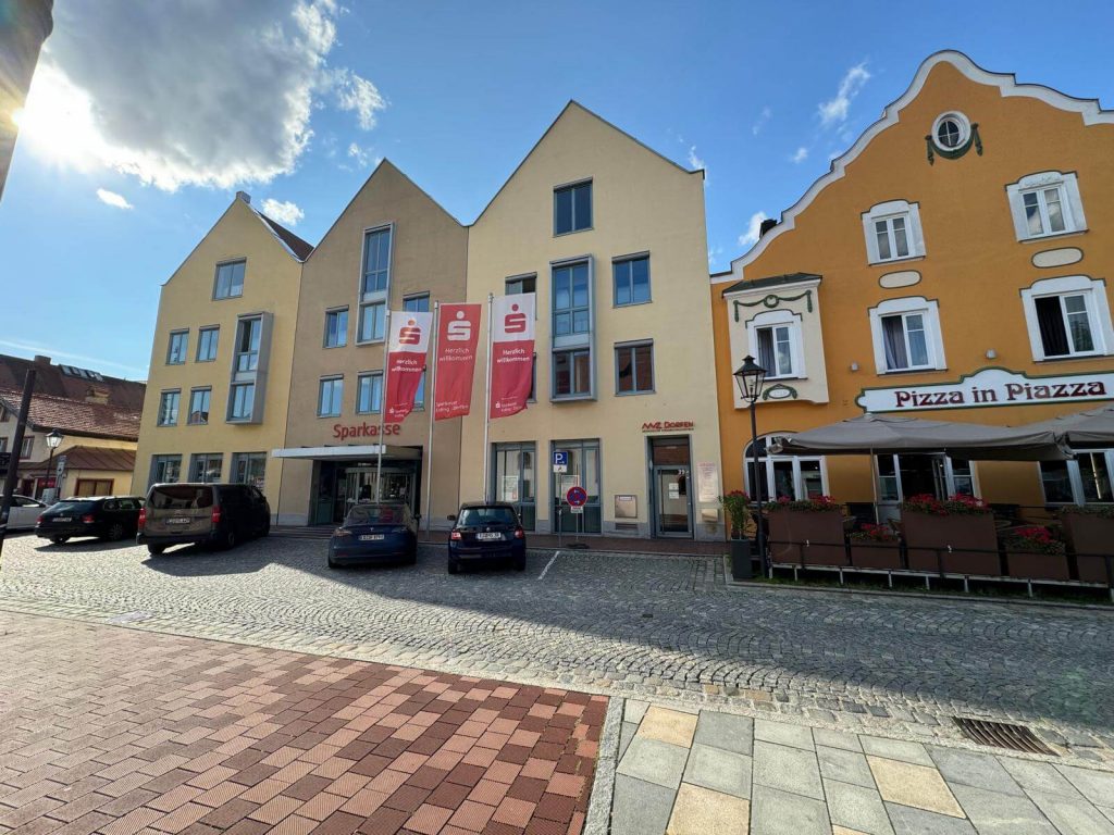 Sie sehen eine Außenansicht eines vierstöckigen schmalen Hauses mit spitzem Giebel. Die Fassade ist sandfarben und über der Tür ist der Schriftzug MVZ Dorfen zu lesen. Links davon befinden sich zwei Häuser identischer Bauart in leicht abgewandelten Farben, rechts davon ein ockerfarbenes Gebäude.