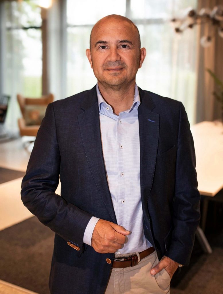 Sie sehen einen Mann mittleren Alters in dunkelblauem Jackett und hellblauem Hemd sowie beiger Hose mit braunem Gürtel. Er trägt eine Glatze und Dreitagebart.