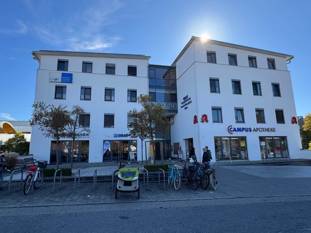 Sie sehen die Außenansicht eines vierstöckigen Gebäudes. Die Fassade ist weiß, das Haus besteht aus zwei Flügeln, die durch einen kleinen verglasten Mittelteil mit Eingang verbunden sind.