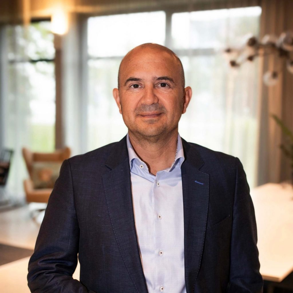 Sie sehen einen Mann mittleren Alters in dunkelblauem Jackett und hellblauem Hemd. Er trägt eine Glatze und Dreitagebart.