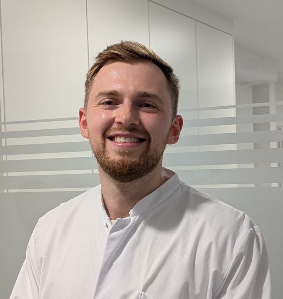 Sie sehen das Portraitfoto eines jungen Mannes in weißem Arztkittel der freundlich lacht. Das dunkelblonde Haar ist kurz und nach links gegelt, er trägt einen Bart.