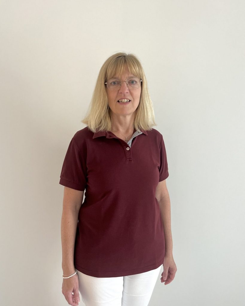 Sie sehen eine Frau mittleren Alters in bordeauxrotem Poloshirt und weißer Hose. Die blonden Haare mit Pony sind schulterlang. SIe trägt eine große ovale Brille mit hellem Gestell.