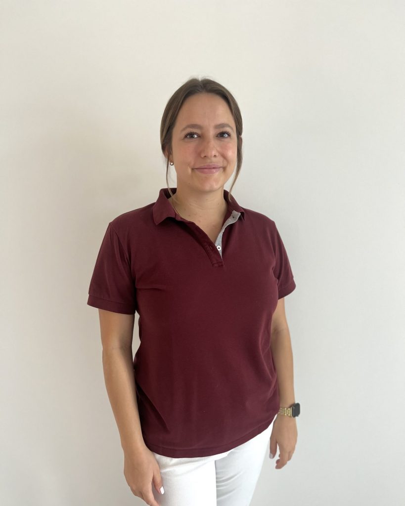 Sie sehen eine lächelnde junge Frau in bordeauxrotem Poloshirt und weißer Hose. Das braune Haar ist zusammengebunden und neben den Schläfen fällt jeweils eine Sträne heraus.