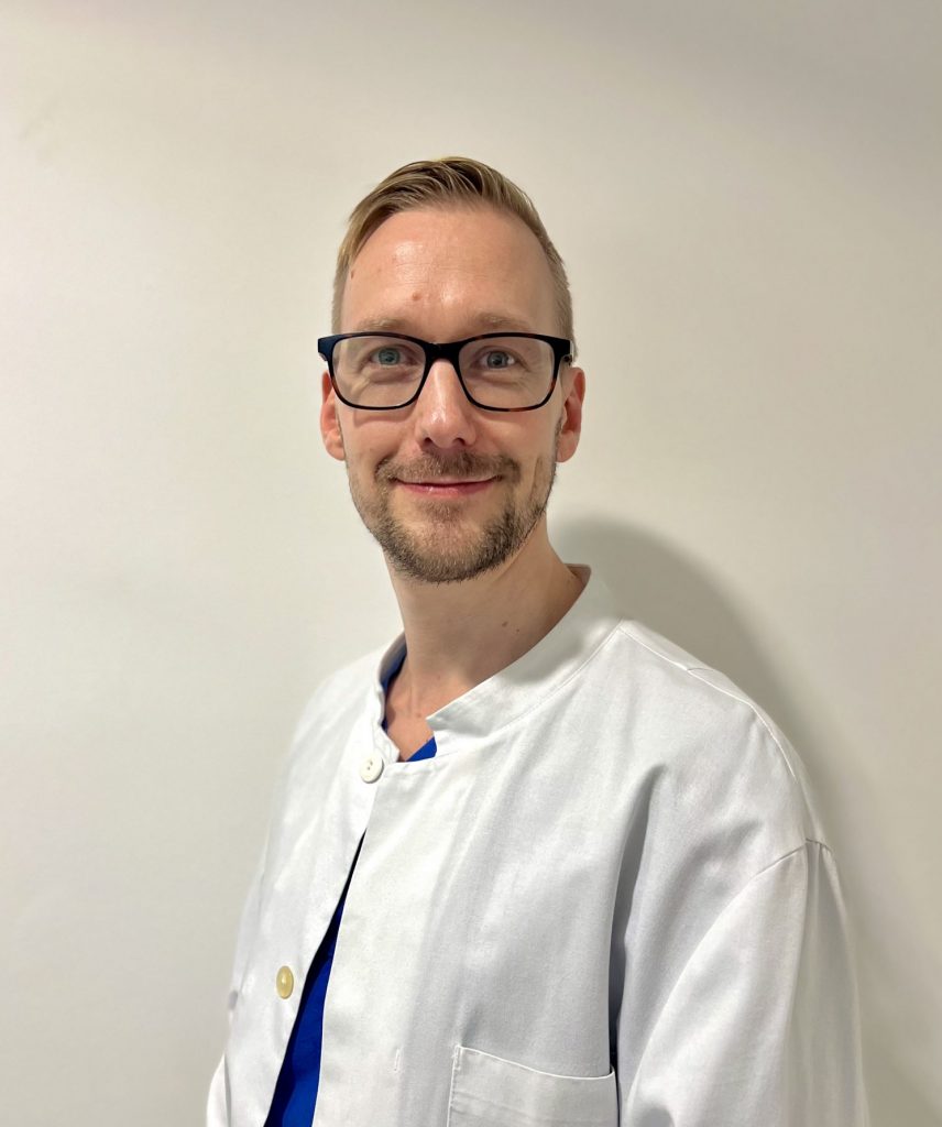 Sie sehen das Portraitfoto eines jungen Mannes in weißem Arztkittel der freundlich lächelt. Das dunkelblonde Haar ist kurz, er trägt einen Bart und Brille.
